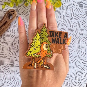 Puede incluir: Una pegatina con un personaje de dibujos animados de Bigfoot frente a dos &aacute;rboles. La pegatina tiene el texto "TAKE A WALK YOU'LL FEEL BETTER" en marr&oacute;n y blanco. El Bigfoot es naranja y los &aacute;rboles son amarillos y verdes.