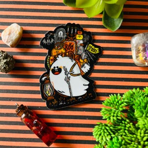 May include: A Halloween-themed sticker featuring a cartoon ghost holding a lantern and a staff. The sticker has a black border and includes the words "Boo" and "Spooky". The background is orange and black striped.
