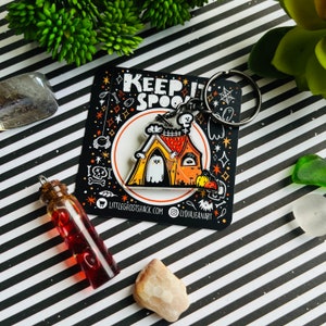 以下が含まれることがあります： ハロウィーンをテーマにしたキーホルダー。家のデザイン、お化け、そして「Keep It Spooky」の文字が特徴です。キーホルダーは黒色で、オレンジ、黄色、白のアクセントがあります。赤い液体が入った小さなガラス瓶も画像にあります。