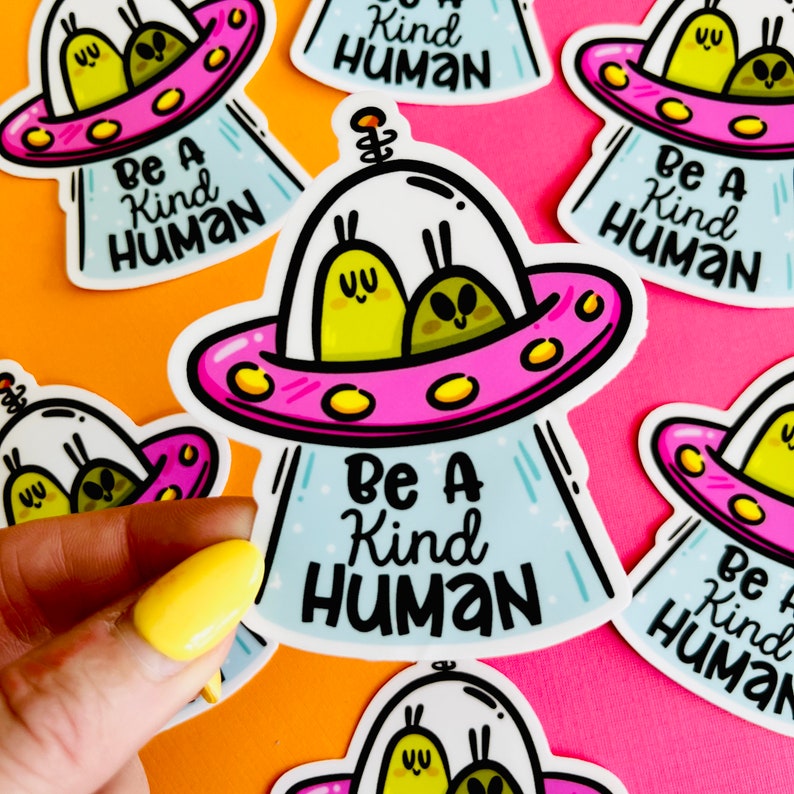 K&ouml;nnte beinhalten: Ein Set mit f&uuml;nf Aufklebern, die ein Cartoon-Alien-Raumschiff mit zwei Aliens darin zeigen. Das Raumschiff ist rosa und wei&szlig; mit gelben Akzenten. Der Text auf den Aufklebern lautet "Be a Kind Human".