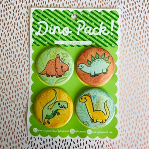 Puede incluir: Un conjunto de cuatro botones redondos, coloridos, con temática de dinosaurios. Cada botón presenta una especie de dinosaurio diferente, incluyendo un triceratops, un estegosaurio, un tiranosaurio rex y un braquiosaurio. Los botones están dispuestos en forma de cuadrado sobre un fondo verde con estrellas blancas. El texto "Dino Pack!" está impreso en blanco sobre un fondo a rayas verde y blanco.
