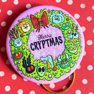 Puede incluir: Un bot&oacute;n redondo rosa con un dise&ntilde;o de corona verde con monstruos de dibujos animados y el texto "Merry Cryptmas". La corona incluye un lazo rojo y copos de nieve. Un lazo dorado est&aacute; unido a la parte posterior.