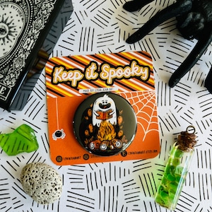 May include: A black and white button with a ghost reading a book by a campfire. The button has the text "Keep it Spooky" and "Made by Lydia Jean Art" on it.