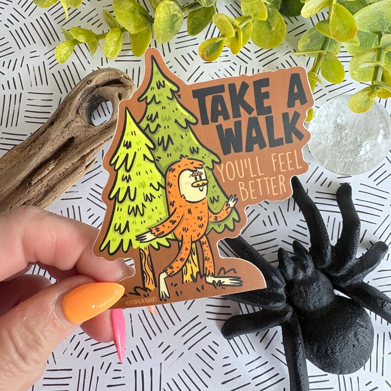 Puede incluir: Una pegatina con un personaje de dibujos animados de Bigfoot frente a dos pinos verdes. La pegatina tiene el texto "TAKE A WALK YOU'LL FEEL BETTER" en naranja y marr&oacute;n. La pegatina est&aacute; sujeta por una persona con esmalte de u&ntilde;as naranja y rosa.