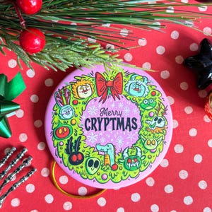Puede incluir: Un adorno navide&ntilde;o redondo y rosa con un dise&ntilde;o de corona verde y amarilla. La corona presenta monstruos de dibujos animados y el texto "Merry Cryptmas" en blanco. Una cinta roja est&aacute; en la parte superior.