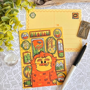 May include: A yellow postcard with a cartoon illustration of a furry orange monster surrounded by framed pictures of a forest, mountains, and a UFO. The postcard has a space for a message and an address. The text "TO:" and "CYCLOJEANART.ETSY.COM" are printed on the postcard.