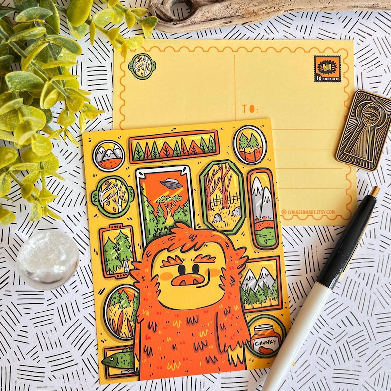 May include: A yellow postcard with a cartoon illustration of a furry orange monster surrounded by framed pictures of a forest, mountains, and a UFO. The postcard has a space for a message and an address. The text "TO:" and "CYCLOJEANART.ETSY.COM" are printed on the postcard.