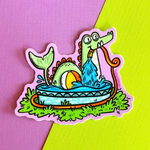 May include: A die-cut sticker featuring a cartoon alligator in a small blue swimming pool. The alligator holds an orange garden hose spraying water into the pool. A beach ball floats in the water, and a green tail rises from the pool. The text LYDIAJEANART is printed vertically on the right side.