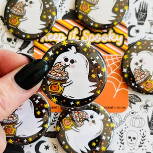 May include: A set of 1 inch button badges featuring a cartoon ghost holding a pumpkin spice latte. The ghost is white with black eyes and a pink blush. The latte is orange and brown with whipped cream and sprinkles. The background is black with yellow stars. The text "Keep it Spooky" is on a yellow and orange striped background.