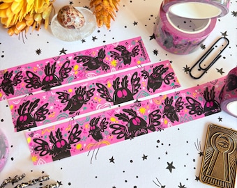 May include: Pink decorative washi tape featuring a repeating pattern of black moth-like creatures with wings, stars, and colorful accents. Rolls of tape and strips are displayed. The tape is ideal for scrapbooking and crafts.