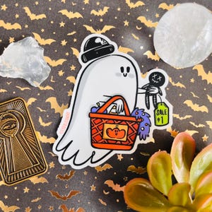 Puede incluir: Un fantasma blanco con un sombrero negro y sosteniendo una cesta de cuadros rojos y blancos con una calabaza en ella. El fantasma también sostiene una pequeña araña negra con una etiqueta que dice "SALE #1".