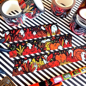 Pu&ograve; includere: Rotoli e strisce di nastro washi decorativo a tema Halloween. Il nastro presenta uno sfondo nero con illustrazioni di fantasmi, zucche e fogliame autunnale in rosso, arancione e giallo. &Egrave; visibile anche una penna con illustrazioni a tema Halloween.