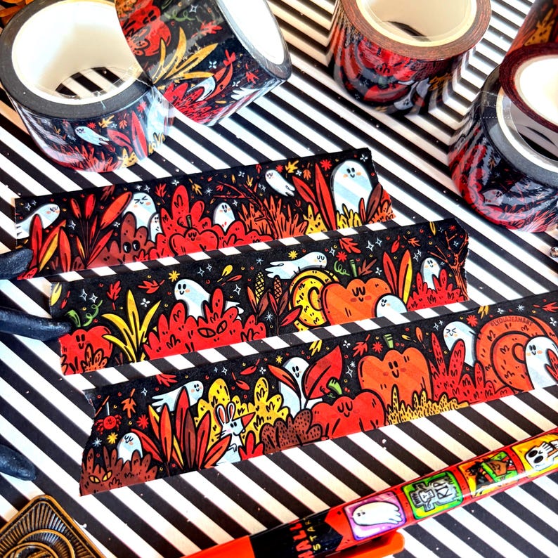 Pu&ograve; includere: Rotoli e strisce di nastro washi decorativo a tema Halloween. Il nastro presenta uno sfondo nero con illustrazioni di fantasmi, zucche e fogliame autunnale in rosso, arancione e giallo. &Egrave; visibile anche una penna con illustrazioni a tema Halloween.