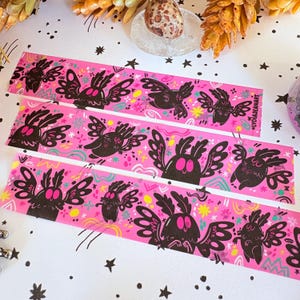 Puede incluir: Tres tiras de cinta decorativa de color rosa brillante con un patr&oacute;n repetitivo de criaturas negras similares a polillas con grandes ojos y alas rosas. El dise&ntilde;o incluye peque&ntilde;as formas abstractas amarillas y verde azulado.