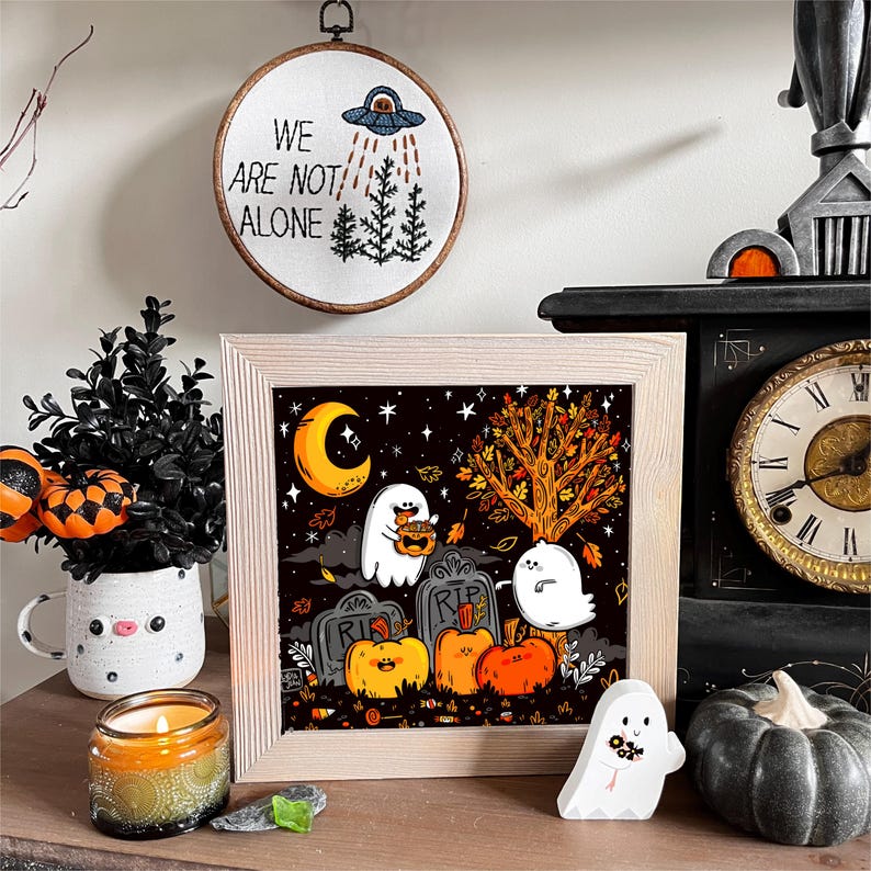 May include: A framed illustration of two ghosts in a graveyard setting. The ghosts are white and have black eyes. They are holding pumpkins and sweets. The background is a dark night sky with stars and a crescent moon. The text "RIP" is on two of the tombstones. The illustration is surrounded by a light wood frame.
