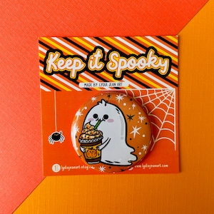 May include: A round button featuring a cartoon ghost enjoying a pumpkin spice latte. The ghost is white with a cute face and is holding the latte with both hands. The background is orange with white stars and a spider web. The text "Keep it Spooky" is at the top of the button and "Made by Lydia Jean Art" is below the ghost. The button is on a card with a black and yellow striped background.