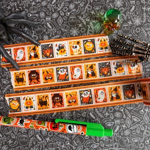 Puede incluir: Tres tiras de cinta adhesiva decorativa y un bol&iacute;grafo retr&aacute;ctil con un patr&oacute;n repetitivo de cuadrados naranjas y negros. Cada cuadrado contiene una ilustraci&oacute;n de dibujos animados de personajes de Halloween como fantasmas, calabazas, murci&eacute;lagos y gatos.