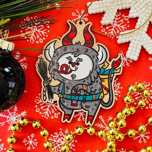 May include: A wooden Christmas ornament shaped like a cartoon monster with red horns, a blue backpack, and a red tongue sticking out. The ornament is painted in a whimsical style and has a festive feel.