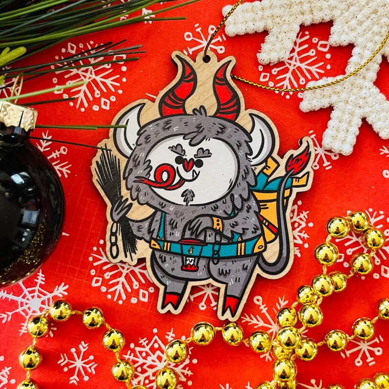 May include: A wooden Christmas ornament shaped like a cartoon monster with red horns, a blue backpack, and a red tongue sticking out. The ornament is painted in a whimsical style and has a festive feel.