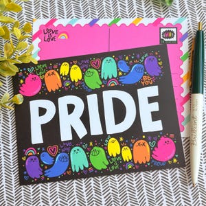 May include: A black postcard with the word "PRIDE" in large white letters, surrounded by colorful cartoon ghosts and rainbows. The postcard has the text "LOVE IS LOVE" and "BE YOU". A pink postcard and a pen are in the background.