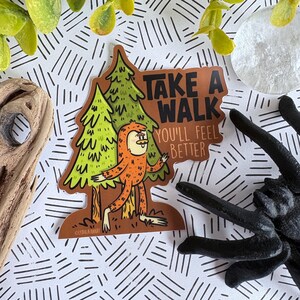Puede incluir: Una pegatina con un personaje de dibujos animados de Bigfoot de pie frente a dos pinos verdes. La pegatina tiene el texto "TAKE A WALK YOU'LL FEEL BETTER" en una pancarta marr&oacute;n. La pegatina est&aacute; sobre un fondo blanco con l&iacute;neas negras.