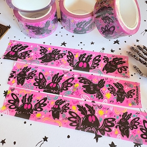 Puede incluir: Tres rollos de cinta adhesiva rosa con un patr&oacute;n repetitivo de criaturas negras parecidas a polillas con ojos y alas grandes. La cinta muestra formas abstractas coloridas, estrellas y el texto LYDIAJEANART impreso en negro en el borde.