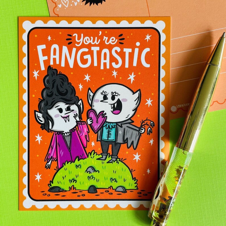 May include: A greeting card with an orange background and the text "You're Fangtastic." The card features cartoon vampires, one holding a heart. A gold pen is in the background.
