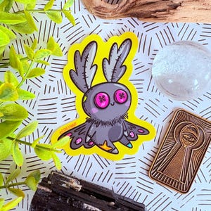 Puede incluir: Una pegatina caprichosa con una criatura de dibujos animados con cuerpo gris, ojos de botón rosa y detalles amarillos. La pegatina tiene un borde amarillo y el texto "LYDIA JEANART". Otros elementos incluyen una esfera transparente y un objeto dorado en forma de ojo de cerradura.