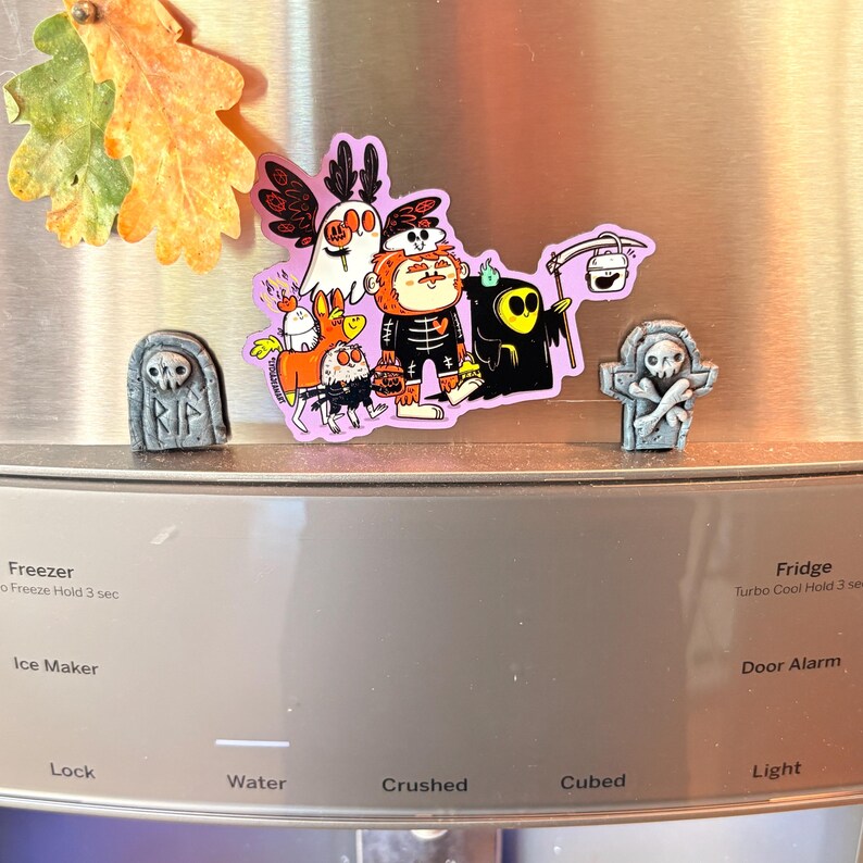 May include: A collection of Halloween-themed magnets, including a tombstone, a group of cartoon characters, and a grim reaper. The magnets are affixed to a stainless steel refrigerator door. The colors are vibrant, with purple, orange, and black dominating the designs.