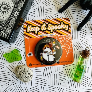 May include: A black and white button with a ghost wearing a witch hat and holding a lantern. The button has the text "Keep it Spooky" and "Made by Lydia Jean Art" on it. The button is on a card with an orange and yellow striped background.