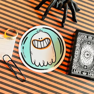 Puede incluir: Una pegatina redonda con un fantasma blanco con una gran sonrisa y dientes naranjas sobre un fondo azul verdoso. La pegatina tiene un contorno negro y el texto "@jvnwilliamart" en la esquina inferior izquierda.