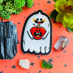 Puede incluir: Un fantasma blanco con una cara amigable sostiene una calabaza roja con una cara sonriente. El fantasma tiene ojos amarillos y una pequeña boca. La calabaza tiene un tallo verde y un tallo marrón.