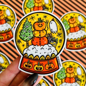 May include: A round, clear snow globe sticker with a festive Halloween theme. Inside the globe, a smiling pumpkin wearing a hat sits on a bed of white snow. The globe is surrounded by snowflakes and other festive details.