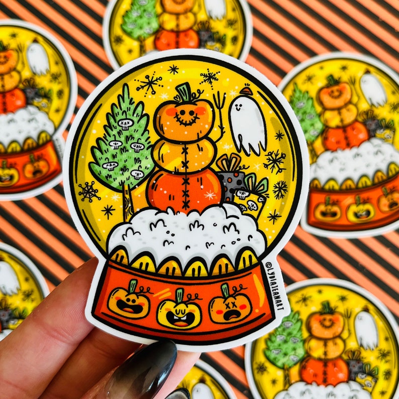 May include: A round, clear snow globe sticker with a festive Halloween theme. Inside the globe, a smiling pumpkin wearing a hat sits on a bed of white snow. The globe is surrounded by snowflakes and other festive details.