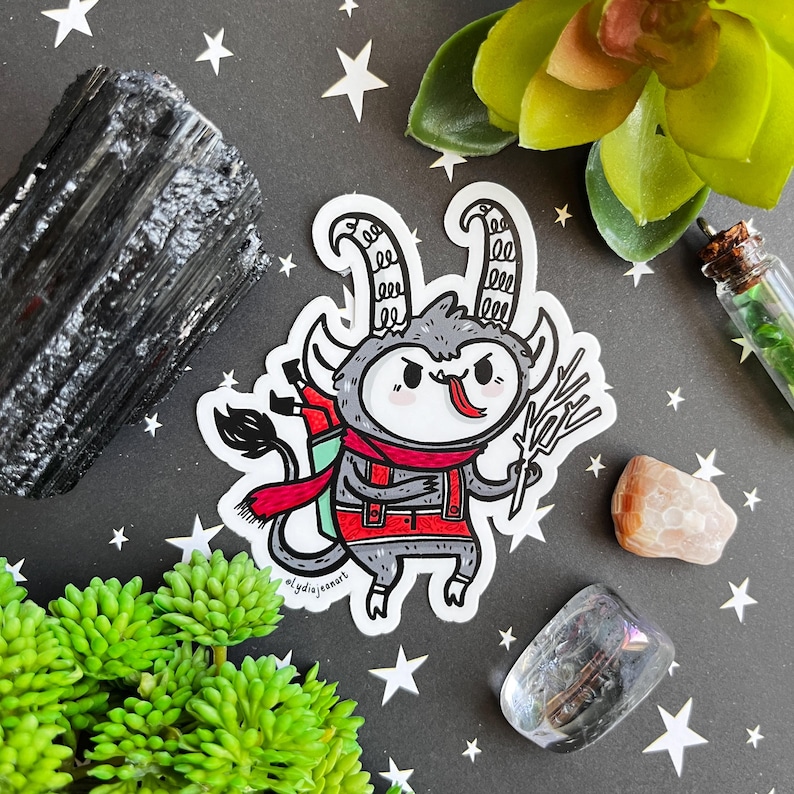 May include: A sticker featuring a cartoon illustration of a gray and white creature with red horns, wearing a red scarf and red overalls, holding a branch in its right hand. The creature has a red tongue sticking out and a mischievous expression. The sticker is on a black background with stars and crystals.