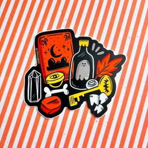 May include: A sticker with a collection of mystical objects. An orange book with a crescent moon and stars, a bottle with a ghost, a yellow key, a crystal, a bone, and an orange box are displayed on a striped background.