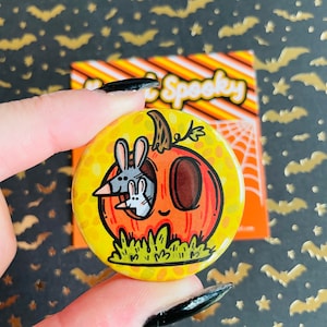 May include: A round button with a cartoon illustration of a pumpkin with a smiling face. Two small white rabbits are peeking out of the pumpkin's opening. The pumpkin is orange with a green stem and leaves. The background is a yellow and orange polka dot pattern.