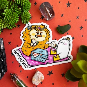 May include: A sticker featuring a cartoon illustration of a furry orange creature with a flower crown, a white ghost-like creature, and a picnic basket on a pink and white blanket. The sticker is on a red background with white stars.