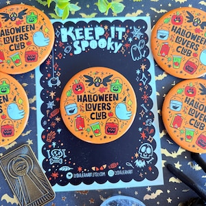 May include: A collection of orange Halloween-themed badges featuring the text "Halloween Lovers Club" and spooky illustrations. A black card with the words "Keep It Spooky" and decorative elements is in the center. The items are on a patterned surface.
