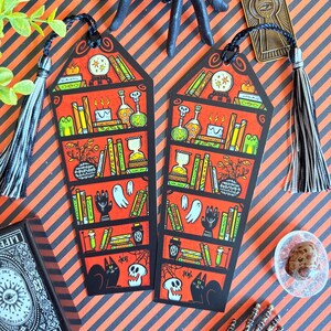 以下が含まれることがあります： 黒いタッセルが付いた、ハロウィーンをテーマにした2つのブックマーク。各ブックマークは、ポーションボトル、本、幽霊、黒猫のイラストが描かれた赤い本棚のデザインが特徴です。背景はオレンジと黒のストライプです。