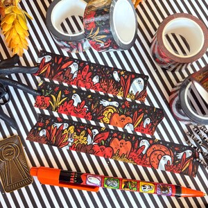 Pu&ograve; includere: Un assortimento di articoli di cancelleria a tema Halloween su una superficie a righe bianche e nere. Include rotoli di nastro decorativo con disegni floreali e fantasia, segnalibri fantasia e una penna arancione con illustrazioni a tema Halloween.