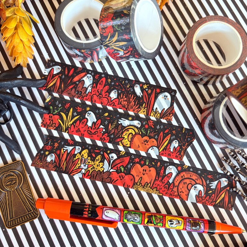 Pu&ograve; includere: Un assortimento di articoli di cancelleria a tema Halloween su una superficie a righe bianche e nere. Include rotoli di nastro decorativo con disegni floreali e fantasia, segnalibri fantasia e una penna arancione con illustrazioni a tema Halloween.