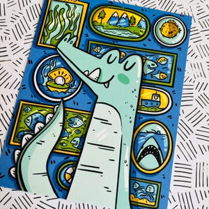 May include: A light blue alligator with a toothy grin is looking at a framed picture of a pearl in a seashell. The alligator is surrounded by other framed pictures of sea life, including fish, a shark, a pufferfish, and a boat.
