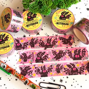 Puede incluir: Varios rollos de cinta washi rosa con una ilustraci&oacute;n repetitiva de Mothman en negro y rosa, con el texto "MYSTERIOUS MOTHMAN" e "ILLUSTRATED BY LYDIA JEAN ART". Tres tiras de cinta est&aacute;n desenrolladas, mostrando el mismo patr&oacute;n gr&aacute;fico.