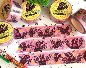 Puede incluir: Varios rollos de cinta washi rosa con una ilustración repetitiva de Mothman en negro y rosa, con el texto "MYSTERIOUS MOTHMAN" e "ILLUSTRATED BY LYDIA JEAN ART". Tres tiras de cinta están desenrolladas, mostrando el mismo patrón gráfico.