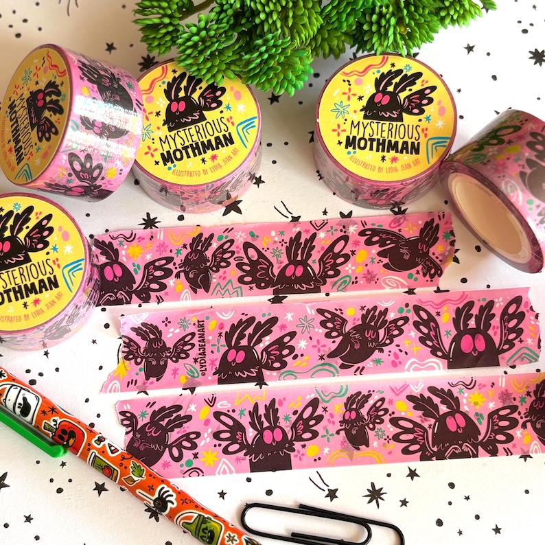 Puede incluir: Varios rollos de cinta washi rosa con una ilustraci&oacute;n repetitiva de Mothman en negro y rosa, con el texto "MYSTERIOUS MOTHMAN" e "ILLUSTRATED BY LYDIA JEAN ART". Tres tiras de cinta est&aacute;n desenrolladas, mostrando el mismo patr&oacute;n gr&aacute;fico.