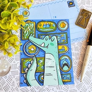 May include: A blue and green cartoon alligator looking at a framed picture of a pearl inside a seashell. The alligator is surrounded by other framed pictures with various sea-themed illustrations, including fish, a shark, a boat, and a jellyfish.