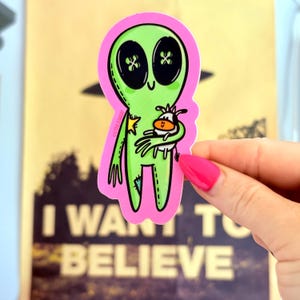 May include: A green alien sticker with large button eyes and a small cow graphic, outlined in black. The alien is holding the cow and is on a pink background. The sticker has the text "I WANT TO BELIEVE".