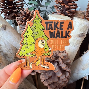 Könnte beinhalten: Ein orangefarbener Aufkleber mit einer Cartoon-Bigfoot-Figur und zwei grünen Kiefern. Der Aufkleber hat den Text "TAKE A WALK YOU'LL FEEL BETTER" in Schwarz. Der Aufkleber wird von einer Hand mit orangefarbenem Nagellack gehalten.