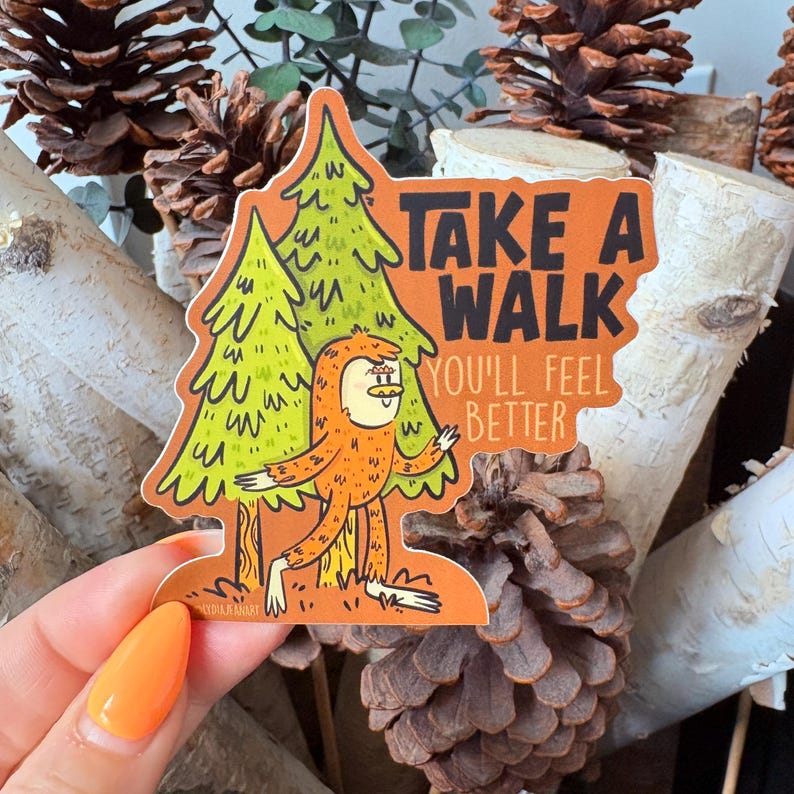 Puede incluir: Una pegatina naranja con un personaje de dibujos animados de Bigfoot y dos pinos verdes. La pegatina tiene el texto "TAKE A WALK YOU'LL FEEL BETTER" en negro. La pegatina est&aacute; sujeta por una mano con esmalte de u&ntilde;as naranja.